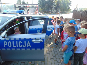 dziewczynka siedząca za kierownicą policyjnego radiowozu, wokół samochodu stoi grupa dzieci