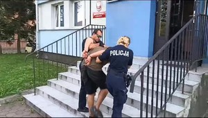 Dwóch umundurowanych policjantów prowadzą zatrzymanego z kajdankami na ręce trzymane z tyłu po schodach do budynku komisariatu