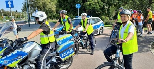 policjant na motocyklu i policjanci na rowerach przy skrzyżowaniu w tle radiowóz i dzieci na jednośladach i rolkach