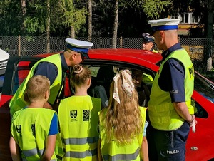 policjanci z uczniami rozmawiają z kierowcą który siedzi w czerwonym samochodzie