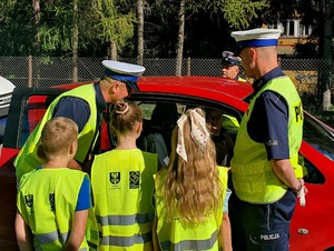 policjanci z uczniami rozmawiają z kierowcą który siedzi w czerwonym samochodzie
