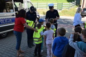 dwoje policjantów rozdaje kamizelki i zawieszki odblaskowe dzieciom, za nimi radiowóz w tle droga