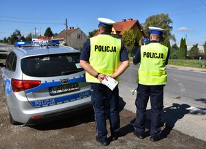 Policjanci nadzorują ruch na drodze. Jeden mierzy prędkość przy użuciu ręcznego miernika prędkości, a drugi trzyma w ręku ulotki