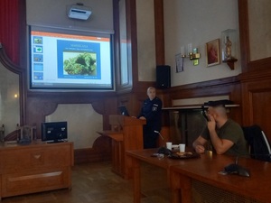 policjantka prowadząca wykład, za nią prezentacja wyświetlona, obok policyjny psycholog siedzący przy stole