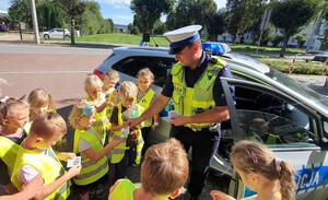 Umundurowany policjant ruchu drogowego rozdaje elementy odblaskowe dzieciom ubranym w kamizelki odblaskowe. osoby stoją obok radiowozu. W tle zabudowania , samochody i drzewa.