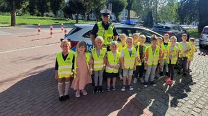 Na zdjęciu widać grupę dzieci ubranych w kamizelki odblaskowe, które stoją przodem, za nimi Umundurowany policjant ruchu drogowego również w kamizelce odblaskowej. W tle budynki, samochody i drzewa.