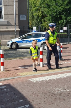 Mała dziewczynka w kamizelce odblaskowej przechodząca przez przejście dla pieszych z umundurowanym policjantem. W tle budynek i oznakowany radiowóz