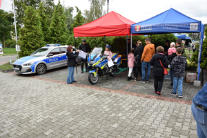 policyjne stoisko, przy nim policjanci i ludzie, obok motor na którym pozuje dziecko, dalej obok zaparkowany radiowóz