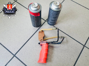 dwie puszki w sprayu z farba połozone obok metalowej procy