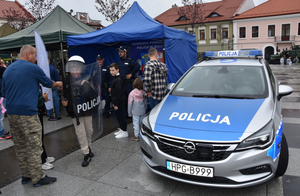 Piknik wojskowy. Jeden z uczestników jest ubrany w elementy policyjnego umundurowania. Trzyma tarczę oraz pałkę.Na zdjęciu widoczny policyjny, oznakowany radiowóz