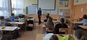 Policjantka stoi przed grupą dzieci. Uczniowie siedzą w szkolnych ławkach.