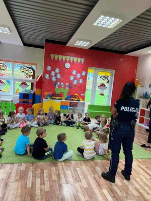 grupa przedszkolaków siedząca na dywanie w kółku, obok stoi policjantka