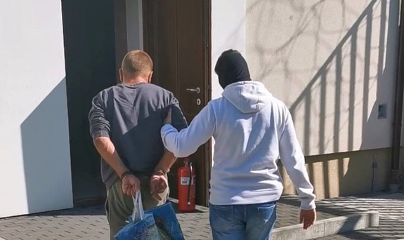 policjant kryminalny prowadzi zatrzymanego z założonymi kajdankami na ręce trzymane z tyłu i wprowadza go do budynku prokuratury