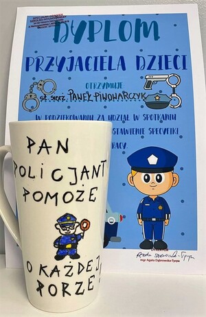 Na zdjęciu widać dyplom dla policjanta za udział w spotkaniu. Przed dyplomem jest postawiony biały kubek z napisem Pan policjant pomoże o każdej porze. Pośrodku jest narysowany policjant
