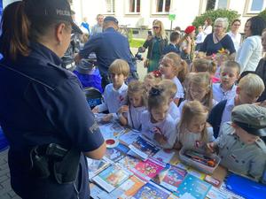 grupa dzieci i dorosłych stojąca przy stoliku z materiałami profilaktycznymi, za stolik stoi policjantka