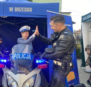 policjant przybijający piątkę chłopcu siedzącego na policyjnym motocyklu, mającego czapkę policyjną na głowie