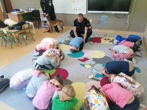 Policjanci tłumaczą w jaki sposób zachowywać sie w trakcie ataku zwierzęcia, dzieci w pozycji żółwia na dywanie