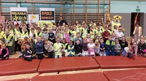 Grupa dzieci stojących i siedzących przodem. Część z nich ma ubrane kamizelki odblaskowe. Za nimi stoi policjant i osoba przebrana za tygrysa. W tle plakaty z napisem Odblaskowa szkoła, Bądź widoczny.