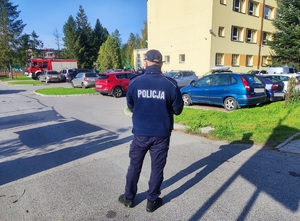 Na drodze wjazdowej do szkoły stoi tyłem umundurowany policjant. W tle budynek szkoły i zaparkowane samochody oraz wóz strażacki.