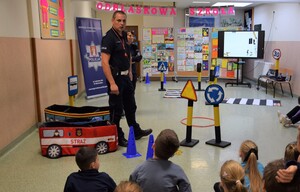 policjant ruchu drogowego podczas prowadzenia prelekcji wraz z policjantką stojącą za nim, przed nimi siedzące na podłodze dzieci, na środku rozłożone miasteczko ze znakami drogowymi