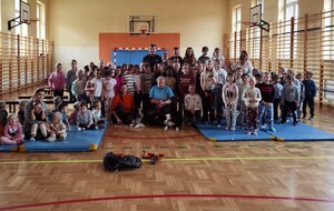 Na zdjęciu widać grupę dzieci siedzących i stojących przodem oraz kobiety w stroju cywilnym. Pośrodku za dziećmi stoi umundurowany policjant. Z przodu psy ratownicze. Osoby sa na sali gimnastycznej.