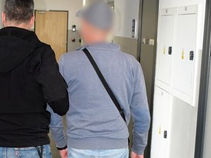 Na zdjęciu po lewej stronie policjant pionu kryminalnego trzymający pod rękę zatzrymanego ubranego w dżinsy, szarą bluzę i szarą czapeczkę z daszkiem. Przepasana ma rówbież czarną małą torbę.