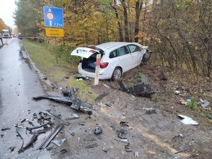 Biały samochód w rowie po kolizji. Auto zniszczone. Na jezdni różne elementy pojazdu. W tle mokra droga i ruch uliczny. Pogoda jesienna.