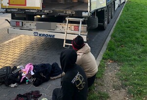 dwóch obcokrajowców siedzących na krawężnikach przed otwartą naczepą, przed sobą mają plecaki