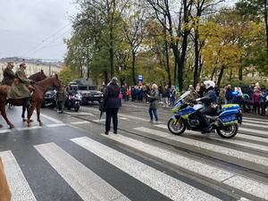 policjant na motocyklu znajdujący się w rejonie przejścia dla pieszych. Na zdjęciu widać także dwóch mężczyzn w umundurowaniu na koniach, pojazdy wojaska oraz zgromadzoną widownię