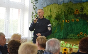 policjant trzymający mikrofon i prowadzący prelekcje skierowaną do seniorów na temat bezpieczeństwa na drodze oraz o zachowaniu ostrzozności w rozmowach telefonicznych z oszustami