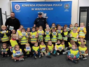 Na tle baneru z napisem Komenda Powiatowa Policji w Wadowicach stoją i siedzą dzieci w kamizelkach odblaskowych. Za nimi umundurowany policjant ruchu drogowego i policjant w stroju cywilnym.