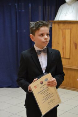 II miejsce Duch
Na zdjęciu stoi chłopiec klasa V, trzyma w ręku dyplom oraz pudełko z nagrodą, w którym znajduje się nagroda. Chłopiec ubrany jest w ciemny garnitur oraz muszkę.