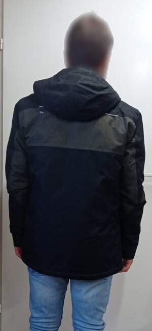 Zatrzymany mężczyzna stojący tyłem na tle białych drzwi. Ubrany w kurtkę koloru ciemnego i niebieskie dżinsy