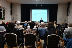 Grupa osób siedząca na krzesłach, osoby są tyłem. Przed nimi stoi umundurowany policjnat, na tle ekranu, na którym jest wyświetlana informacja dotycząca kradzieży i zagubienia dokumentów.