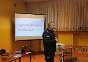 Policjant w sali szkolnej prowadzi prelekcje  w tle rzutnik i stand wyświelacza