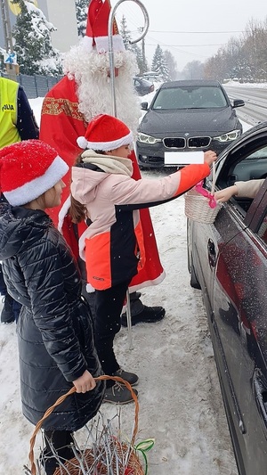 Na zdjęciu widać dwie dziewczynki w czapkach Mikołaja, maja koszyczki. Obok Mikołaj. Po prawej stronie fragment samochodu, którego kierowca zaniera z koszyka cukierek. W tle czarny samochód.