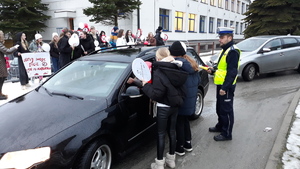 Kilka młodych osób rozmawia z kierowcą pojazdu, obok stoi policjant. W tle stoi grupa młodzieży