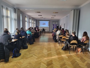 uczniowie siedzący w sali tyłem do zdjęcia, przed nimi policjant i wyświetlona prezentacja oraz baner komendy