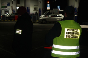 stojący tyłem nieumundurowany policjant z opaska z napisem POLICJA, obok niego stojący tyłem funkcjonariusz ITD