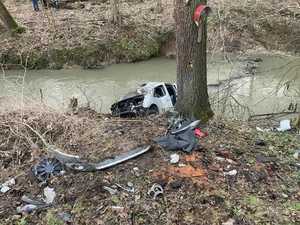 pojazd po wypadku,  leżący na brzegu rzeki, częściowo w niej zanurzony, obok drzewo, obok niego leżące poodrywane części samochodu