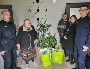 dwóch policjantów umundurowanychz pracownikiem opieki spolecznej, obok nich staruszka i jej syn, na ziemi ułożone siatki z zakupionymi darami dla rodziny