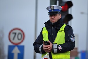 Policjant ruchu drogowego nadzoruje ruch na drodze. W ręku trzyma krótkofalówkę. W tle widać znaki drogowe i sygnalizator nadający czerwone światło