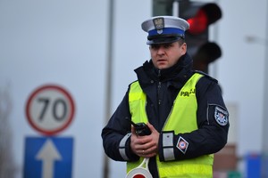 Policjant ruchu drogowego nadzoruje ruch na drodze. W ręku trzyma krótkofalówkę. W tle widać znaki drogowe i sygnalizator nadający czerwone światło