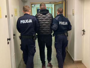 dwóch umundurowanych policjantów prowadzi zatrzmanego mężczyznę przez korytarz policyjnej jednostki