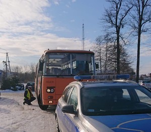 policjant sprawdza stan techniczny podwozia, schylony obok autobusu. Na pierwszym planie radiowóz za nim zaparkowany autobus szkolny koloru pomarańczowego