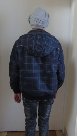 Zatrzymana kobieta stojąca tyłem na tle białych drzwi. Ubrana w niebieskie dżinsy, kurtke granatowa w kratkę i szarą czapkę.