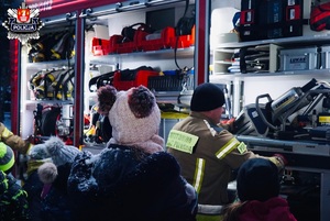 dzieci w czapach i kurtakch zimowych stojace przy bezkowozie strażąckim oglądaja szczypce hydrauliczne prezentowane przez strażaka