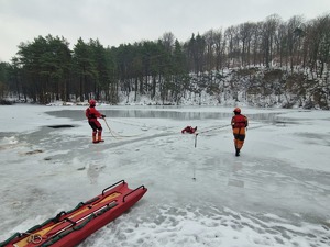 Dwóch strażaków w specjalnych ubraniach ratowniczych stoja w dużej odległości od siebie i za pomoca lin wydobywają poszkodowana osobę spod lodu. W tle zamarznięty staw i krajobraz