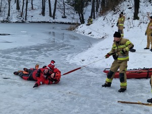 Strażak przy zamarzniętym stawie, ciagnie line, przy której umocowany jest ratonik oraz poszkodowany. Symulacja ratowania człowieka spod lodu.