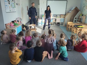 dwoje umundurownych policjantów stojących przed siedzącymi na podłodze przedszkolakami